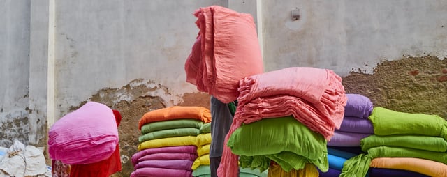 Stacks of brightly colored fabric bundles, including pink, green, orange, purple, and blue, arranged against a weathered concrete wall